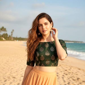 May include: A dark green crop top with a gold floral pattern and a peach skirt. The outfit is worn on a sandy beach with the sea in the background. The top has short sleeves and a boat neckline.