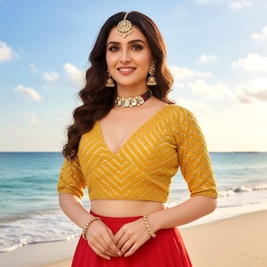 Könnte beinhalten: Eine Frau trägt ein gelbes Crop-Top mit goldenen Akzenten und einen roten Rock. Sie ist mit goldenem Schmuck geschmückt, darunter Ohrringe, eine Halskette und Armbänder. Ein dekorativer Kopfschmuck vervollständigt das Ensemble. Im Hintergrund sind Strand und Meer zu sehen.