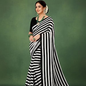 Könnte beinhalten: Ein schwarz-weiß gestreifter Sari mit einer schwarzen Bluse. Der Sari hat kräftige vertikale Streifen. Die Frau trägt grünen Schmuck, darunter eine Halskette, Armbänder und einen Ring. Der Hintergrund ist einfarbig grün.