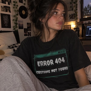 Może przedstawiać: Czarny t-shirt z krótkim rękawem z zieloną, prostokątną grafiką przypominającą okno komputerowe. Grafika zawiera biały napis "ERROR 404" w górnym wierszu oraz "COSTUME NOT FOUND" w dolnym wierszu, wraz z małymi ikonami sterowania oknem.