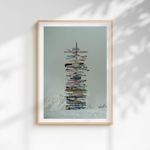 Op de afbeelding: Een ingelijste print met een hoge stapel kleurrijke vintage ski's, gerangschikt als een wegwijzer. De ski's zijn in verschillende kleuren en ontwerpen, met het woord "ISCHGL" zichtbaar bovenaan. De achtergrond toont een besneeuwd berglandschap.