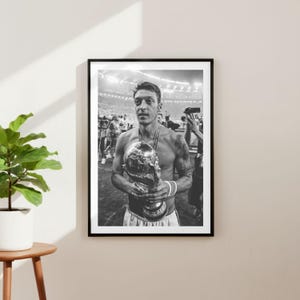 May include: Black and white framed print of a soccer player holding a trophy. The player is shirtless, wearing a medal, and has tattoos on his arm. The background shows a stadium with people.