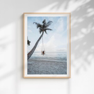 May include: Framed photograph of a tropical beach scene. A person is swinging on a swing attached to a leaning palm tree. The beach has white sand, and the ocean is a light blue color. The sky is a pale blue with some clouds.