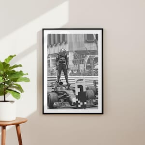 Puede incluir: Impresión enmarcada en blanco y negro de un coche de carreras de Fórmula 1 con un piloto de pie encima. El piloto lleva un traje y un casco de carreras. La impresión se muestra en una pared blanca.