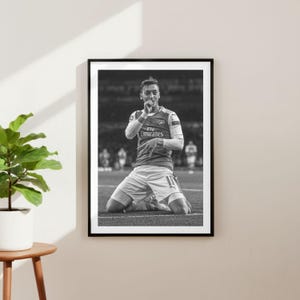 May include: Black and white framed print of a soccer player kneeling on a field, celebrating. The player wears a red and white jersey with the Emirates logo. The image is set against a blurred background of other players and stadium seating.