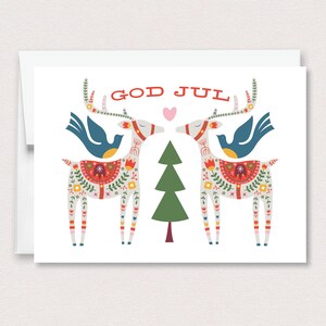 May include: A white greeting card featuring a colorful illustration of two reindeer facing each other with a green pine tree in the center. The reindeer have floral patterns, blue birds on their backs, and a pink heart above. The text reads GOD JUL.