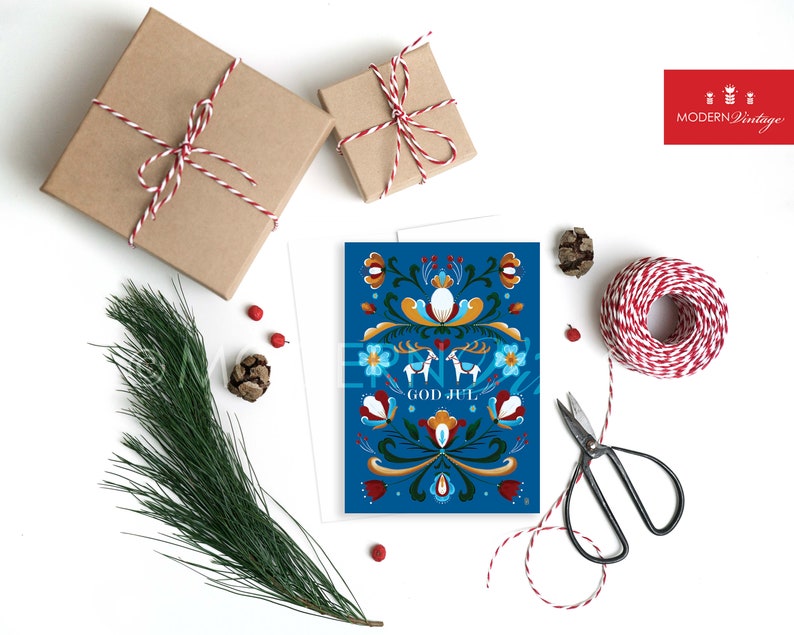 May include: A blue Christmas card with a traditional Scandinavian folk art design featuring a floral pattern, two reindeer, and the text "God Jul".