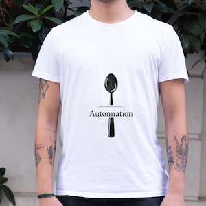 以下が含まれることがあります： 黒いスプーンのグラフィックと下に「Automation」という文字が入った白いTシャツ。シャツは柔らかい素材で作られています。背景には緑の葉と白い壁が含まれています。
