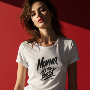 May include: White t-shirt with rolled sleeves featuring the text "Momma the Best" in a black brushstroke font. The shirt is paired with blue jeans against a red background. The Stussy logo is below the text.