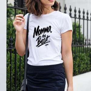 May include: A white t-shirt with the text "Momma the Best" in a black, handwritten style. The shirt is worn with a navy blue skirt and a black shoulder bag. The background includes a black wrought iron fence.