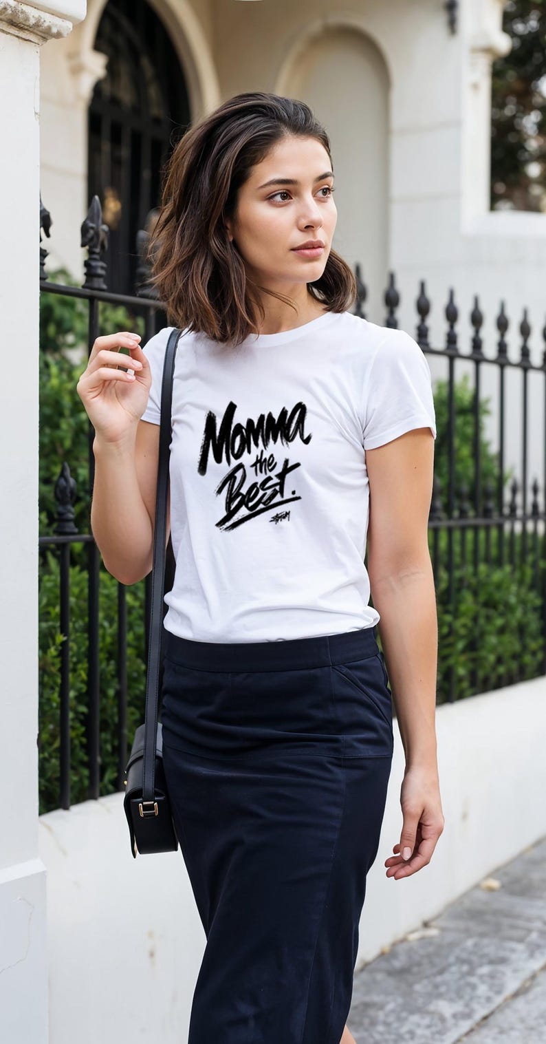 May include: A white t-shirt with the text "Momma the Best" in a black, handwritten style. The shirt is worn with a navy blue skirt and a black shoulder bag. The background includes a black wrought iron fence.