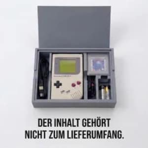Könnte beinhalten: Eine graue, rechteckige Aufbewahrungsbox mit Klappdeckel, die eine beige tragbare Spielkonsole, ein schwarzes Ladekabel, eine Spielkassette und zwei Batterien enthält. Der Text unter der Box lautet: DER INHALT GEHÖRT NICHT ZUM LIEFERUMFANG.
