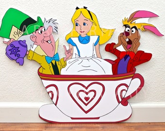 May include: Alice in Wonderland characters, the Mad Hatter, Alice, and the March Hare, are sitting in a large teacup with red and white hearts. The Mad Hatter is holding a purple teapot with a star on it. The March Hare is holding his hands to his face in surprise. Alice is wearing a blue dress and a white apron. The teacup has a gold rim and a red handle.