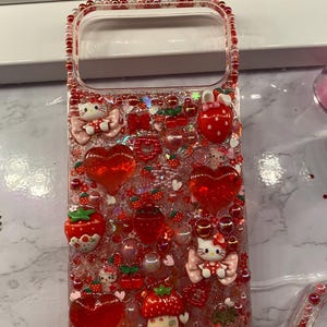 May include: Clear phone case decorated with red glitter, hearts, strawberries, and Hello Kitty figures. The case has a beaded border and various 3D embellishments, creating a playful and decorative design.