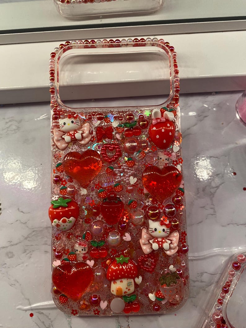 May include: Clear phone case decorated with red glitter, hearts, strawberries, and Hello Kitty figures. The case has a beaded border and various 3D embellishments, creating a playful and decorative design.