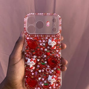 May include: A clear phone case decorated with red hearts, strawberries, and Hello Kitty figures. The case is embellished with small red and pink beads. A small figure is near the camera lenses.