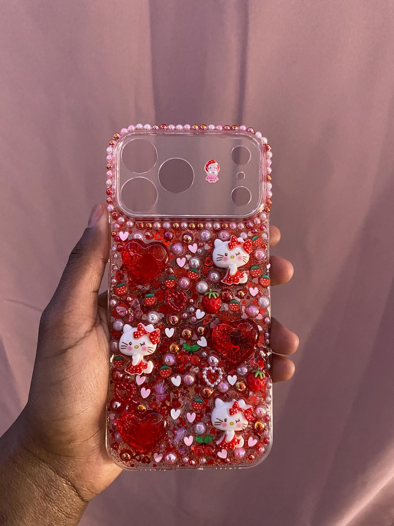 May include: A clear phone case decorated with red hearts, strawberries, and Hello Kitty figures. The case is embellished with small red and pink beads. A small figure is near the camera lenses.