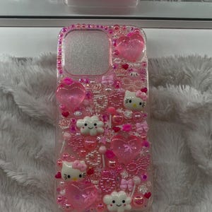 May include: A clear phone case decorated with pink embellishments, including hearts, bows, Hello Kitty characters, and small beads. The case is adorned with a border of pink rhinestones. A matching case is visible in the background.