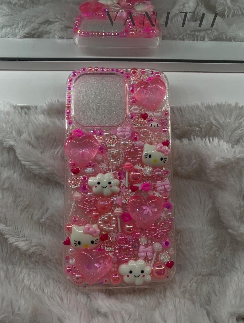 May include: A clear phone case decorated with pink embellishments, including hearts, bows, Hello Kitty characters, and small beads. The case is adorned with a border of pink rhinestones. A matching case is visible in the background.