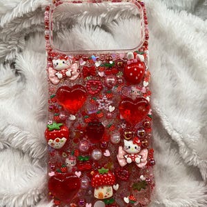 May include: A clear phone case decorated with red and pink embellishments. The case features heart-shaped charms, strawberries, bows, and Hello Kitty figures. The edges are lined with pink beads, creating a whimsical and playful design.