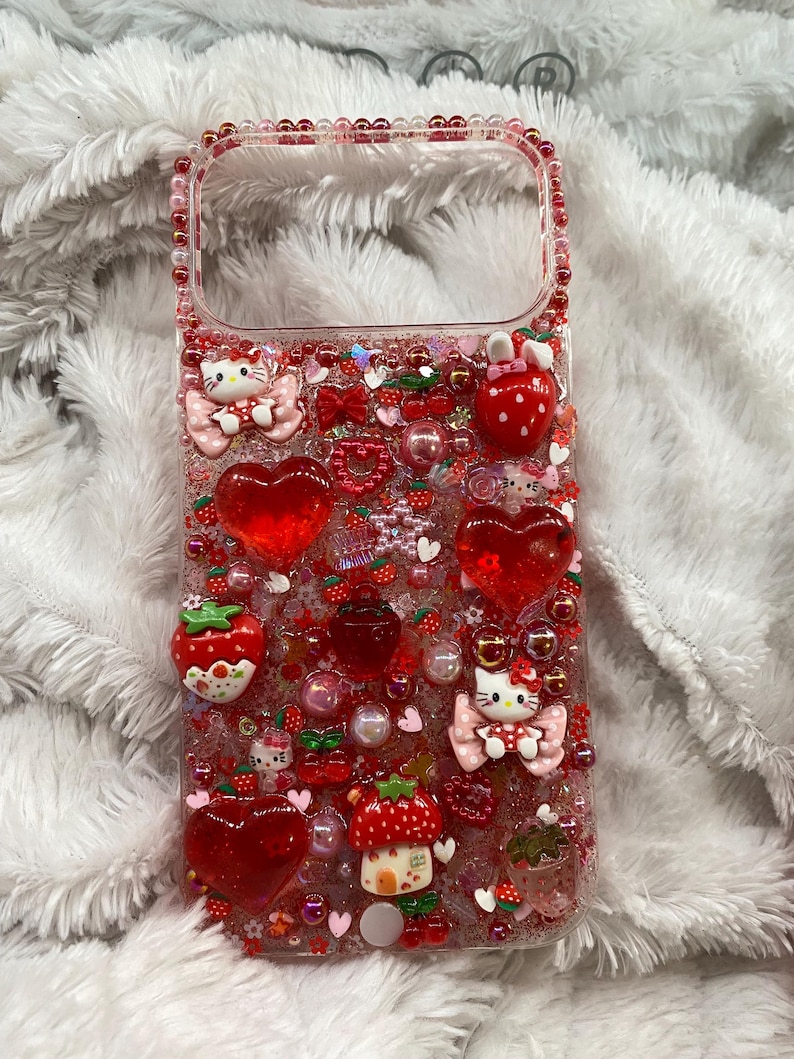 May include: A clear phone case decorated with red and pink embellishments. The case features heart-shaped charms, strawberries, bows, and Hello Kitty figures. The edges are lined with pink beads, creating a whimsical and playful design.