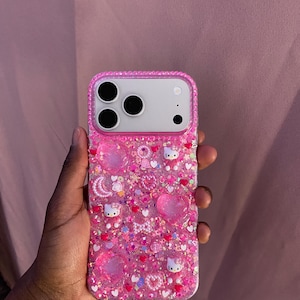 May include: Pink phone case with a glittery, textured surface. The case is adorned with heart-shaped embellishments, small Hello Kitty figures, and various other decorative elements. The case has a clear border with embedded rhinestones.