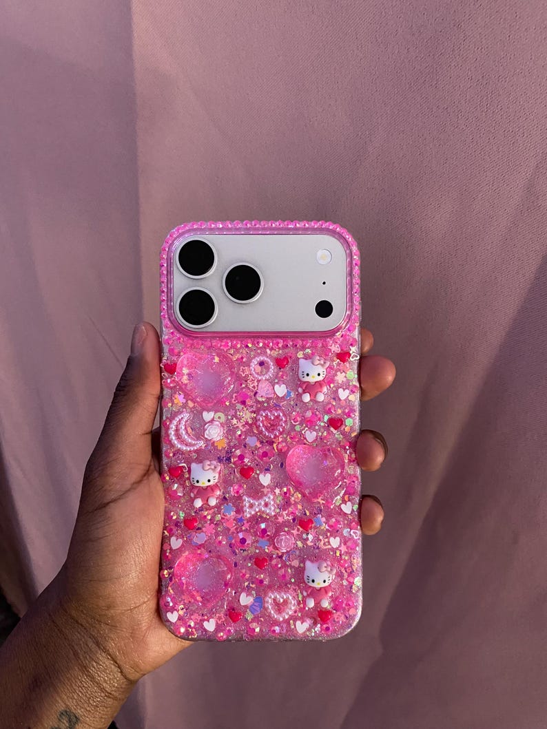 May include: Pink phone case with a glittery, textured surface. The case is adorned with heart-shaped embellishments, small Hello Kitty figures, and various other decorative elements. The case has a clear border with embedded rhinestones.