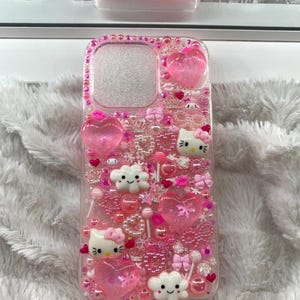 May include: A pink phone case decorated with hearts, bows, beads, and Hello Kitty characters. The case has a clear background and is embellished with various pink and clear accents. The word "VANITII" is visible on a matching case in the background.