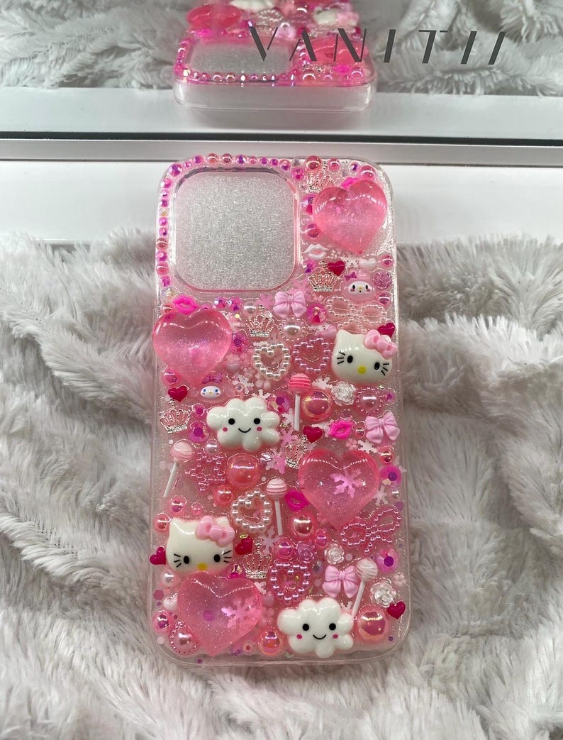 May include: A pink phone case decorated with hearts, bows, beads, and Hello Kitty characters. The case has a clear background and is embellished with various pink and clear accents. The word "VANITII" is visible on a matching case in the background.