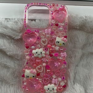 May include: A pink phone case decorated with various embellishments. The case features heart-shaped charms, bows, pearls, and cartoon characters. The case is encrusted with pink rhinestones around the edges.