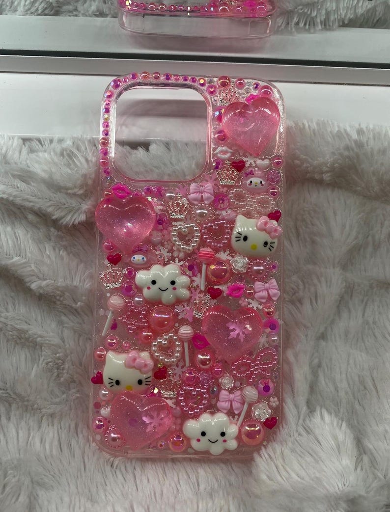 May include: A pink phone case decorated with various embellishments. The case features heart-shaped charms, bows, pearls, and cartoon characters. The case is encrusted with pink rhinestones around the edges.