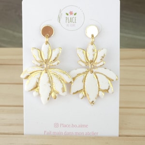 Peut inclure: Une paire de boucles d'oreilles en forme de fleur de lotus blanches et dorées. Les boucles d'oreilles ont un design de pétales superposés avec des accents dorés. Elles sont suspendues à un disque doré. Texte: "Place.bo.aime" et "Fait main dans mon atelier".