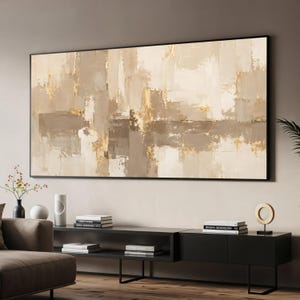 Puede incluir: Una gran pieza de arte mural rectangular que presenta una composición abstracta de pinceladas superpuestas en tonos beige, crema, topo y oro metálico, encerrada en un marco negro fino y exhibida horizontalmente sobre una consola multimedia negra.