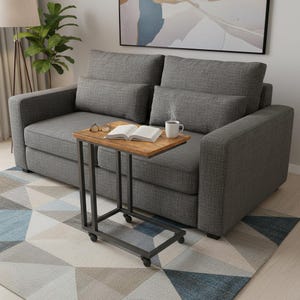 May include: A gray upholstered sofa with two back cushions and two smaller pillows. A small wooden side table with a metal frame and wheels is next to the sofa. The table holds a book, glasses, and a mug of coffee. A patterned rug is on the floor.