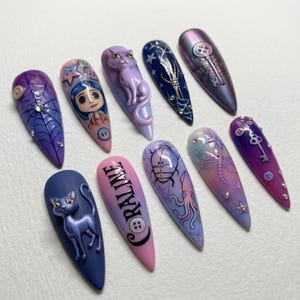 May include: A set of ten long, almond-shaped artificial nails with various designs. The designs include a spiderweb, a cartoon character, a cat, a skeleton, a key, and the word "CORALINE". Colors include purple, blue, pink, and iridescent shades.