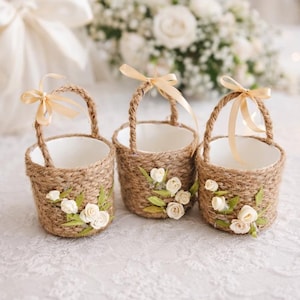 Può includere: Tre piccoli cestini intrecciati con manici e interni color crema. I cestini sono adornati con elementi floreali color crema e legati con nastri dorati. Lo sfondo presenta fiori bianchi e illuminazione soffusa.