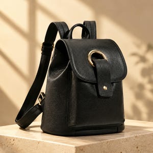 Puede incluir: Una mochila negra con superficie texturizada, que cuenta con un cierre de solapa asegurado por un anillo de metal dorado y un pequeño remache. La bolsa incluye correas ajustables para los hombros, un asa superior y una silueta estructurada.