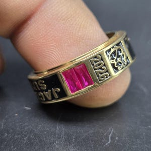 May include: A gold-toned ring with a rectangular, magenta-colored stone. The ring has black enamel sections with the inscription "JAC SU" and the year "2026". Decorative patterns are etched on the ring's surface.