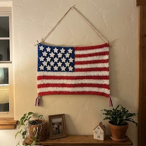 May include: A rectangular crochet wall hanging featuring the American flag design with red, white, and blue yarn. The blue canton contains white star appliqués, and the flag is suspended by a light-colored cord with small tassels at the bottom corners.