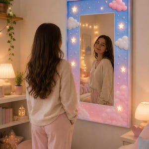 May include: A rectangular wall mirror featuring a light blue frame decorated with three-dimensional pink and white clouds and illuminated stars. The mirror surface reflects a room interior, while the frame itself emits a soft, warm glow.