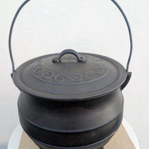 May include: A black cast iron cauldron with a rounded body, three small feet, and a matching lid featuring a raised swirl pattern. The pot includes a thin, arched metal handle attached to the sides, providing a sturdy grip for lifting.
