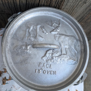 May include: A silver-colored cast iron Dutch oven lid with a deer and fawn relief design. The lid has a handle and the text "MACA 13" OVEN". The lid is approximately 13 inches in diameter.