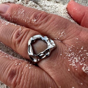 Könnte beinhalten: Ein silberfarbener Ring mit einer organischen, unregelmäßigen Form. Der Ring hat eine strukturierte Oberfläche mit einer großen, offenen Mitte. Der Ring wird an einem Finger getragen, auf einem sandigen Untergrund.