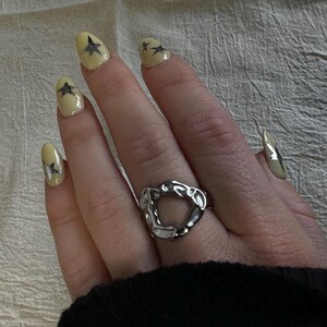 Könnte beinhalten: Ein silberner Ring mit unregelmäßigem, organischem Design wird an einer Hand mit gelbem Nagellack getragen. Die Nägel sind mit schwarzen Sterndesigns verziert. Der Ring ist der Mittelpunkt und zeigt eine moderne, abstrakte Ästhetik.