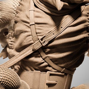May include: A detailed sculpture of a creature wearing a textured fabric outfit with leather straps and a belt. The creature's skin has a scale-like pattern, and the overall color palette is a muted brown.