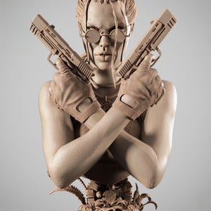 May include: A detailed bust of a character from Tomb Raider, holding two pistols. The figure wears round glasses and fingerless gloves. The base of the bust features the words "TOMB RAIDER". The sculpture is a neutral beige color.