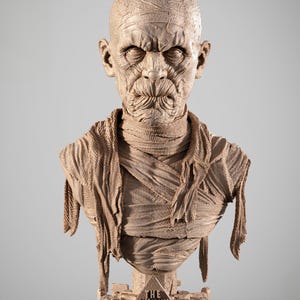 May include: A detailed bust of a mummy with a stern expression, wrapped in textured bandages. The bust is light brown and features the text "THE MUMMY" on the base, along with additional hieroglyphic text. The sculpture is a detailed representation of the classic movie monster.
