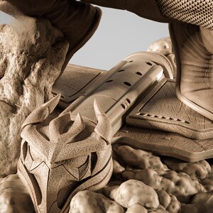 May include: A detailed, sepia-toned sculpture of a stylized helmet or mask, featuring sharp angles and a textured surface. The design includes pointed horns and a central, angular face. The piece is surrounded by a rocky, uneven terrain, suggesting a fantasy or sci-fi theme.