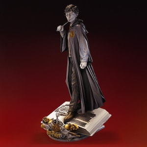 May include: A detailed figurine of Harry Potter, wearing glasses and holding a wand, stands atop an open book. He is dressed in a long gray robe with a gold emblem. The base features the words "Harry Potter".