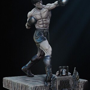 Può includere: Statua grigia di Rocky Balboa in posa da pugile, con guantoni e pantaloncini neri. La figura muscolosa è su una piattaforma. Una targa alla base recita "Rocky Balboa". La statua ha dettagli neri.